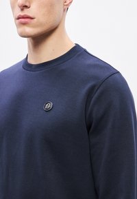 Tmavomodrá bavlnená mikina s okrúhlym výstrihom a logom na hrudi, hladkou textúrou a priliehavým strihom.