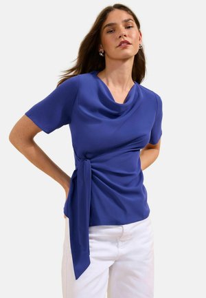 Frau trägt eine royalblaue kurzärmelige Bluse mit drapiertem Halsausschnitt und Seitenschleife, kombiniert mit weißen Hosen, steht mit den Händen in der Hüfte.