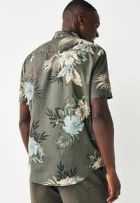 Camisa de manga corta en tela verde oscuro con grandes patrones florales en beis y azul, con un fondo texturizado y un cuello estándar.