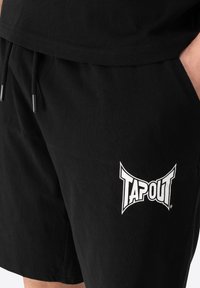 Musta puuvillashortsit, joissa on naru vyötäröllä, sivutaskut ja valkoinen "TAPOUT"-logo vasemmassa jalassa.