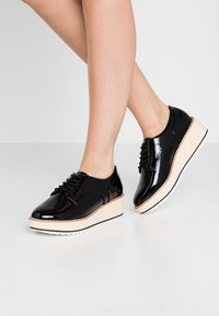 Chaussures compensées en cuir verni noir avec une semelle intermédiaire tissée beige et une semelle extérieure en caoutchouc blanc, dotées d'un design à lacets et d'un bout arrondi.