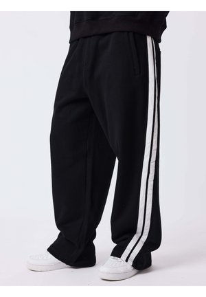 Personne portant un pantalon noir ample avec des bandes blanches sur les côtés et des baskets blanches, debout sur un fond clair uni.