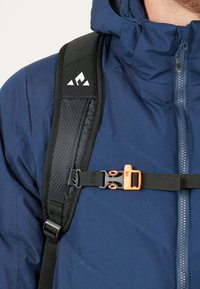 Mörkblå ryggsäck med texturerade svarta remmar, ett orange spänne och en vit logotyp på remmen, med en blixtlåsförsedd jacka delvis synlig.