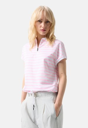 Blondynka ubrana w biały krótki rękaw z różowymi paskami i białe spodnie z sznurkiem, stojąca z rękami w kieszeniach.