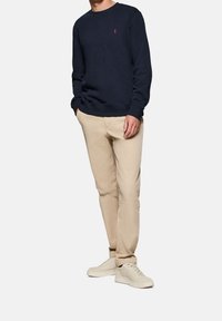 Mörkblå sweatshirt med ribbat halsringning och ärmslut, tillsammans med ljusbruna chinos och beige sneakers. Sweatshirten har en liten röd logotyp.