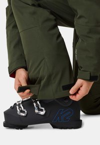 But narciarski z logo K2, w matowej czerni, ze srebrnymi klamrami i teksturowanym uchwytem. Nogawka zielonych spodni jest dopasowana za pomocą paska na rzep.