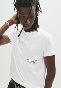 Hvid crew-neck t-shirt lavet af bomuld, med broderet tekst "STWD" i sort og orange, båret med lagdelte sølv kædeder.