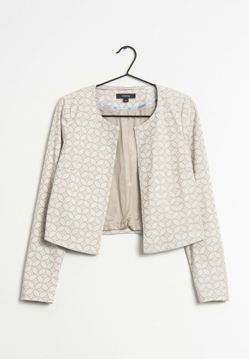 Veste courte beige à manches longues avec motif floral circulaire, suspendue ouverte sur un cintre noir sur fond blanc.
