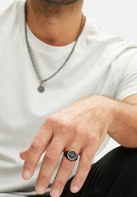 Zwarte metalen ring met een ronde, gepatterneerde schijf, met een geometrisch ontwerp. Ge dragen aan een hand, ter aanvulling van een zilveren kettingketting.