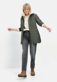 Olijfgroene gewatteerde jas met een capuchon, zijzakken en ritsluiting. Gepaard met grijze jeans en bruin veterschoenen tot de enkels.