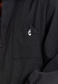 Camisa de tela acanalada negra con un bolsillo delantero que presenta una esquina doblada y un pequeño corazón bordado en blanco con un detalle de bucle.