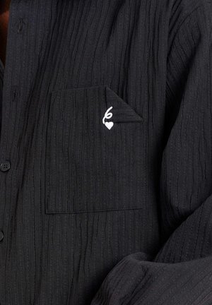 Zwarte ribstof shirt met een voorzak met een gevouwen hoek en een klein wit geborduurd hartje en lusdetail.