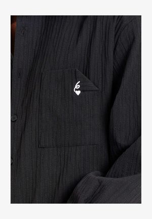 Schwarzes, geripptes Stoffhemd mit einer Fronttasche, die eine gefaltete Ecke und ein kleines weißes, gesticktes Herz mit Schlaufen-Detaillierung aufweist.