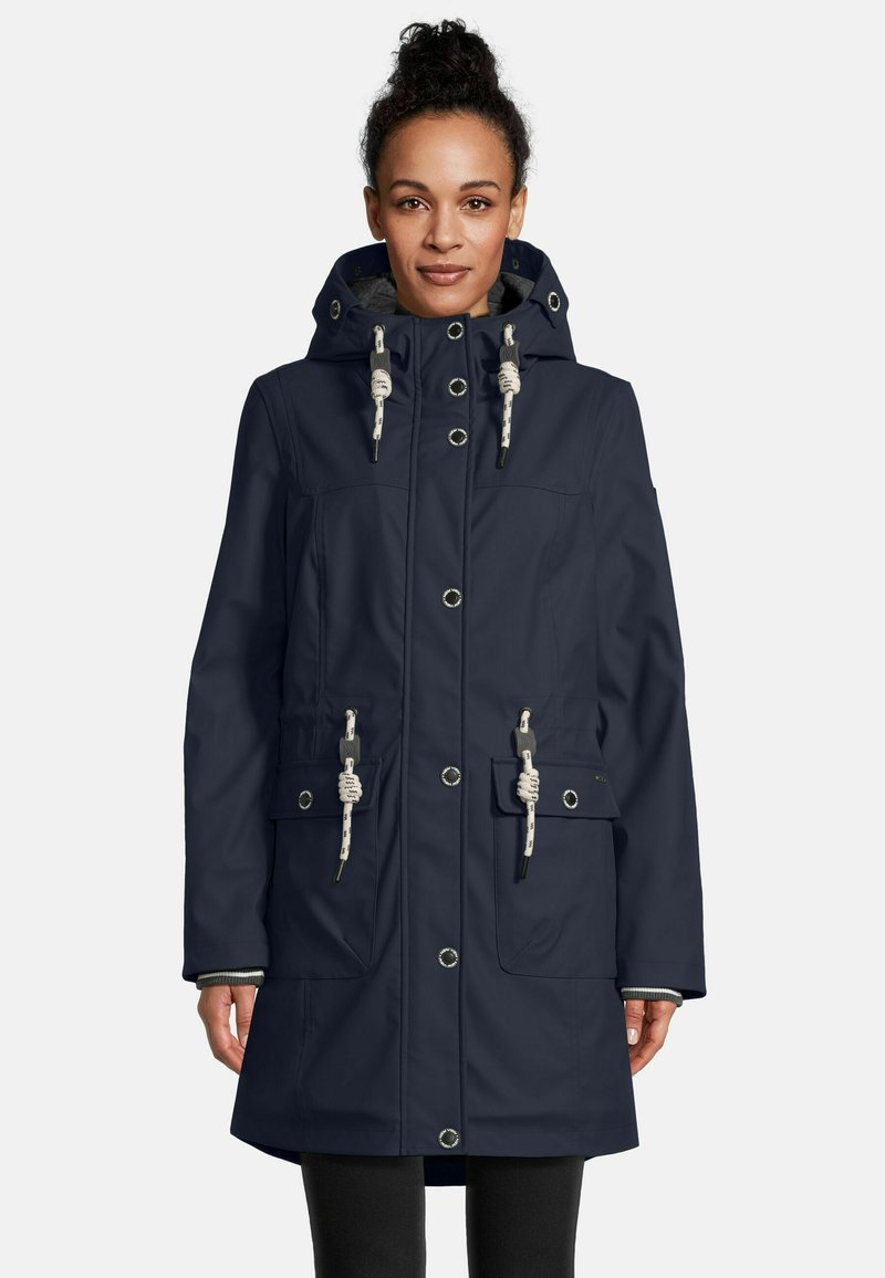 Vrouw in een marineblauwe gewatteerde parka met witte touwsluitingen en voorzakken, staand tegen een plain witte achtergrond.