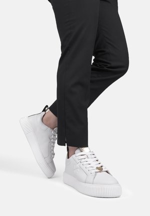 Sneaker bianchi con una texture in pelle liscia, lacci piatti, dettagli dorati e una suola spessa in gomma bianca, abbinati a pantaloni neri a taglio slim.