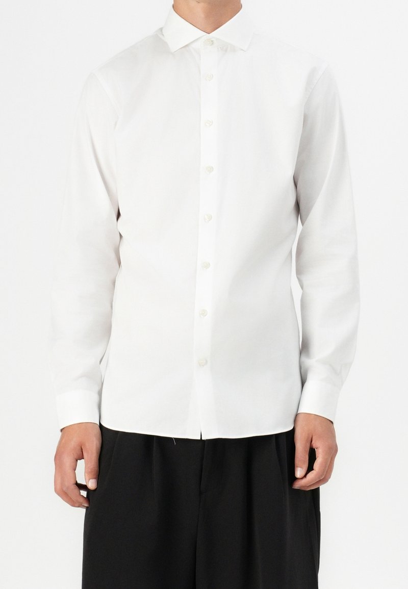 Homme portant une chemise blanche ajustée à manches longues boutonnée avec un col classique et un pantalon noir à jambes larges.