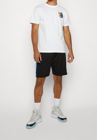 Vit bomull t-shirt med en flerfärgad grafik på vänster bröst, med svarta shorts som har blå sidostripes och ljusa sneakers.