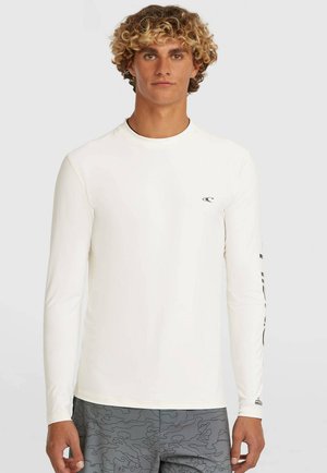 Jonge man met krullend haar, gekleed in een wit shirt met lange mouwen en grijze korte broek met patroon, staand tegen een effen lichte achtergrond.