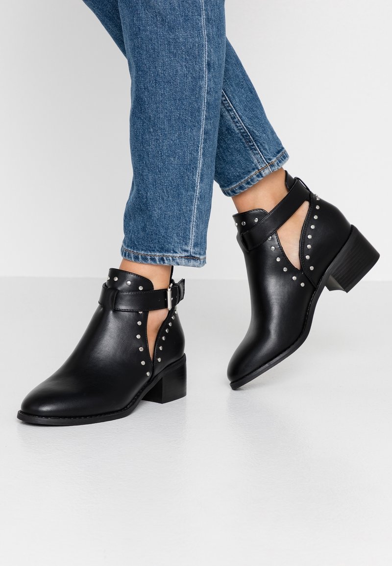 Botas de cuero negras hasta el tobillo con tachuelas plateadas y recortes laterales, llevadas con jeans azules frente a un fondo blanco.