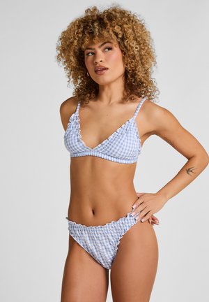 Vrouw met krullend haar, gekleed in een blauw-witte geruite bikini, staand met één hand op de heup tegen een effen achtergrond.