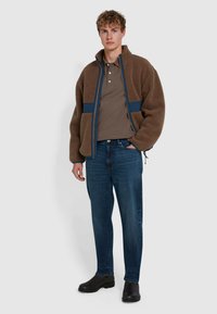 Braune Fleecejacke mit blauen Akzenten und Reißverschluss, kombiniert mit einem braunen Poloshirt und blauen Jeans, abgerundet mit schwarzen Schuhen.