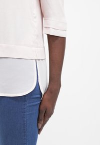 Blusa rosa chiaro con maniche a tre quarti, sovrapposta a uno strato inferiore bianco. Orlo dritto, tessuto morbido e testurizzato. Abbinata a jeans blu.