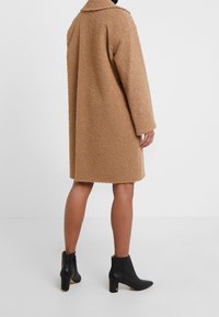 Abrigo de textura esponjosa color marrón canela con cuello, mangas tres cuartos y un corte holgado, combinado con botas de tobillo negras con tacón.