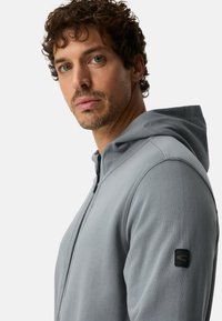 Lichtgrijze hoodie met ritssluiting, gemaakt van zacht materiaal, voorzien van een capuchon en een zwart logo op de linkermouw, met een getailleerd ontwerp.