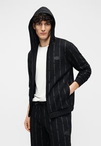 Sudadera con capucha negra con cremallera, hecha de tela suave, con un patrón de logotipo repetido, bolsillos delanteros y capucha con cordón. Combinada con una camisa blanca.