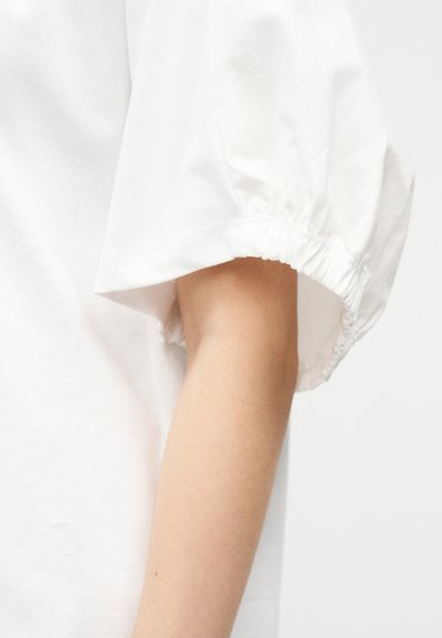 Χέρι που φοράει λευκό πουκάμισο με χαλαρή, σούρα κοντομάνικη λεπτομέρεια σε φωτεινό φόντο.