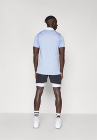 Polo de manga corta en azul claro con cuello blanco, combinado con pantalones cortos negros y calcetas blancas a rayas. Modelo de espaldas, mostrando el ajuste de la prenda.