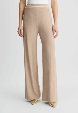 Kobieta nosząca beżowe, szerokie, dzianinowe spodnie z paskiem w paski oraz złote buty z wydłużonym noskiem, stojąca na jednolitym tle.