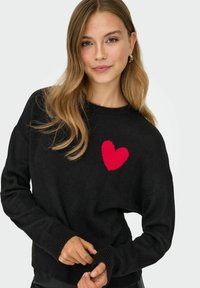 Maglione nero a maglia con un design a forma di cuore rosso sul petto, dotato di polsini a coste e una vestibilità rilassata. Texture morbida e design informale.
