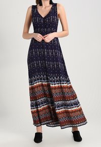 Mouwloze V-hals maxi-jurk met marineblauwe en ingewikkelde meerkleurige geometrische patronen, gedragen met zwarte gesloten schoenen.