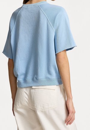 Persoon met een lichtblauw kortarmtruitje met ruwe randen en ecru hoge taillebroek, gezien van achteren tegen een effen achtergrond.