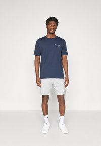 Marineblaues kurzärmeliges T-Shirt mit Logo auf der Brust, kombiniert mit hellgrauen Shorts. Weiße Sneakers und Socken runden das Outfit ab.