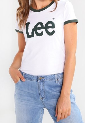 Vrouw draagt een wit T-shirt met donkergroene biezen en het "Lee"-logo, gecombineerd met lichtblauwe spijkerbroek, met één hand in de zak.