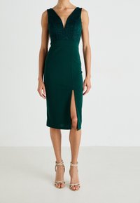 Robe midi sans manches vert foncé avec corsage en dentelle, décolleté en V profond, fente latérale, portée avec des sandales à talons hauts à brides sur un fond neutre.