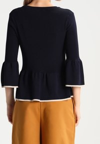 Granatowy top z baskinką, rękawy typu dzwonek o długości do łokcia oraz białe wykończenie wzdłuż dekoltu i brzegu. Gładka dzianina.