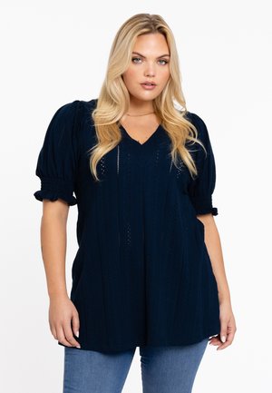 Top en tricot bleu marine avec un col en V, des manches courtes et bouffantes, et un motif texturé comportant des œillets. Coupe décontractée avec des poches sur les coutures latérales.