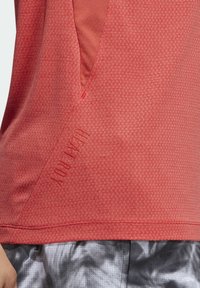 Rød tekstureret atletisk skjorte med mesh-design og et logo på siden, der har en let sideslids for ventilation.