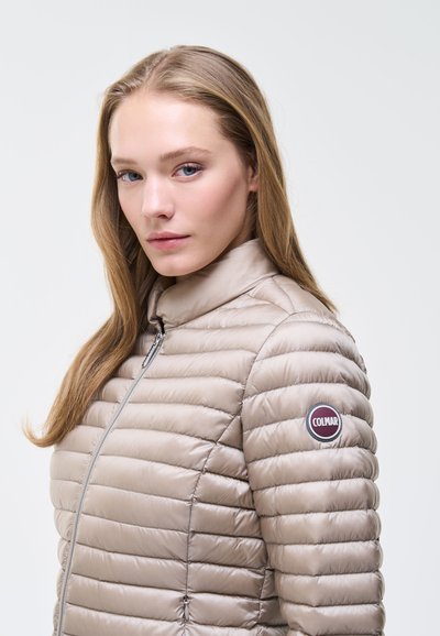 Femme aux longs cheveux blonds portant une veste matelassée beige avec un patch logo Colmar sur le haut du bras, sur fond blanc uni.