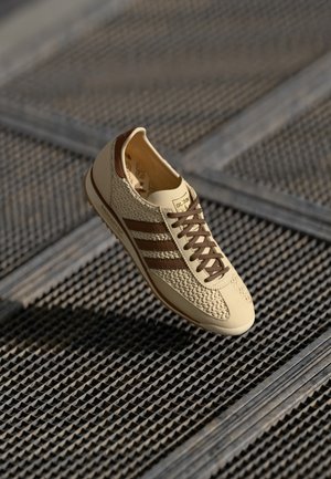Beżowy sneaker z brązowymi paskami i sznurówkami, pleciona tekstura po bokach, unoszący się nad metalową kratownicą podłogi.