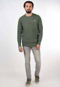 Green sweatshirt with a textured fabric, crew neck, and a small yellow logo on the chest, paired with light grey jeans and green sneakers.