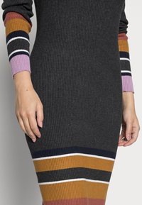 Robe en maille gris foncé avec texture côtelée ; manches longues ornées de rayures multicolores en moutarde, rose, bleu marine et blanc.