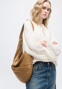 Νεαρή γυναίκα με κυματιστά ξανθά μαλλιά φοράει λευκό πουκάμισο, μπλε τζιν και κρατά μια μεγάλη καφέ δερμάτινη τσάντα ώμου, στέκεται με σταυρωμένα τα χέρια.