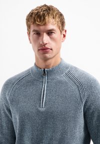 Lichtgrijze ritstrui met hoge hals, geribbelde textuur en gestreepte details op de schouders. Heeft een korte rits bij de hals.