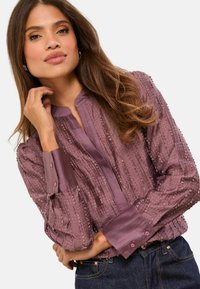 Lila blúz függőleges texturált csíkokkal, álló gallérral, gombos mandzsettával és enyhén bő szabással, sötét farmerrel párosítva.