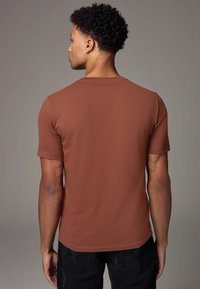 Tricou scurt de culoare maro, din material neted, cu guler rotund și un design ajustat, asortat cu pantaloni închisi la culoare.