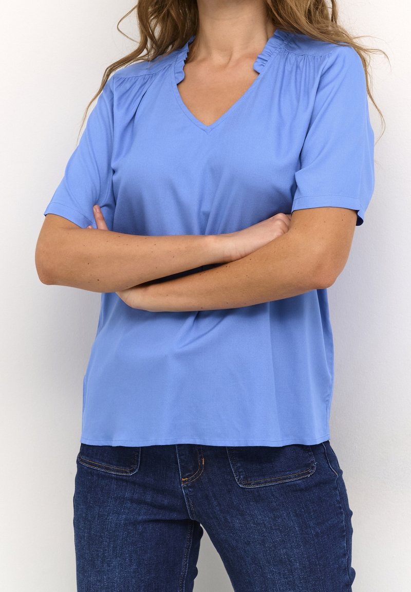 Haut bleu clair à manches courtes avec un col en V et des détails à volants sur le col. Fabriqué en tissu doux, avec une coupe décontractée et une texture lisse.